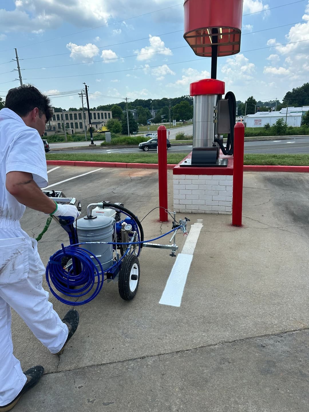 Person in white overalls using a spray painter on a parking lot line near a gas station.