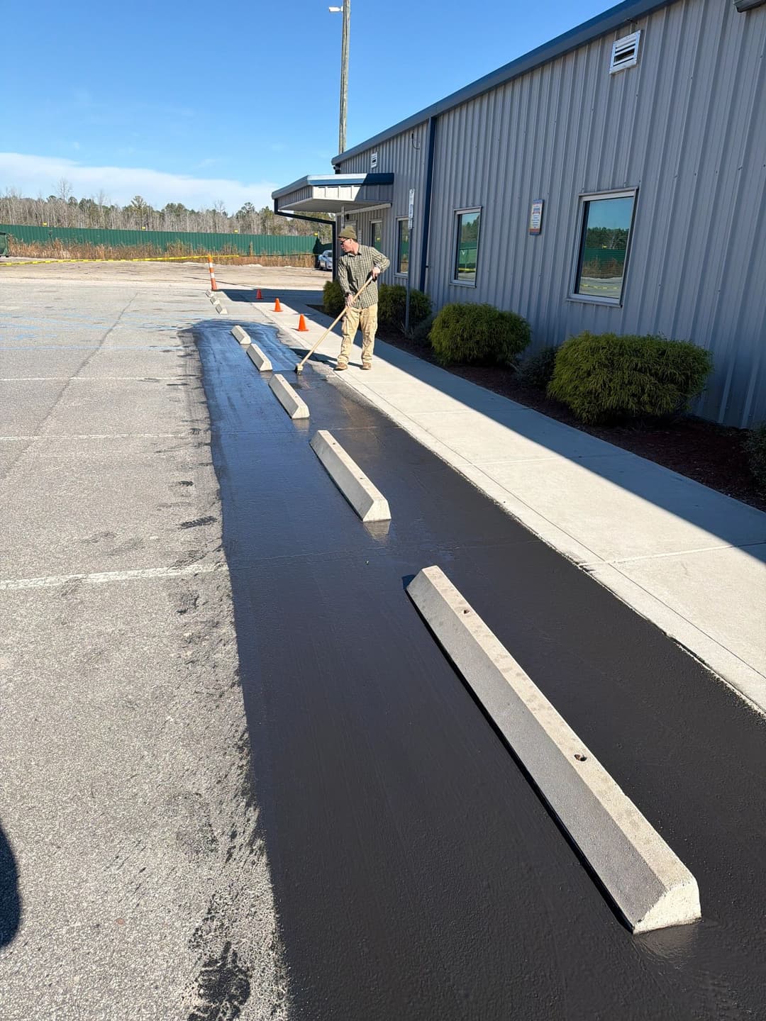 Worker smoothing freshly poured asphalt in a parking lot beside a metal building.